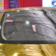 金色のクルマの運転席にはイーロン・マスクのマスク（お面）